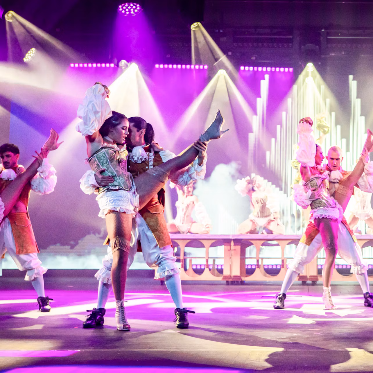 Performers in elaborate costumes dance on a brightly lit stage with colorful lights and a decorative backdrop.