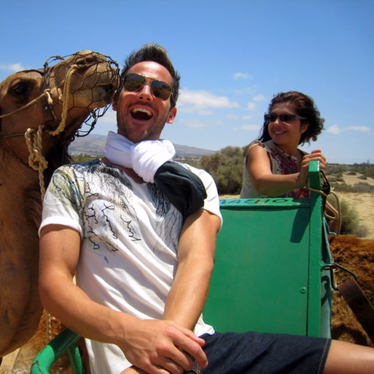 A man laughs while a nearby camel nuzzles his face. A woman smiles in the background, seated on a green cart.