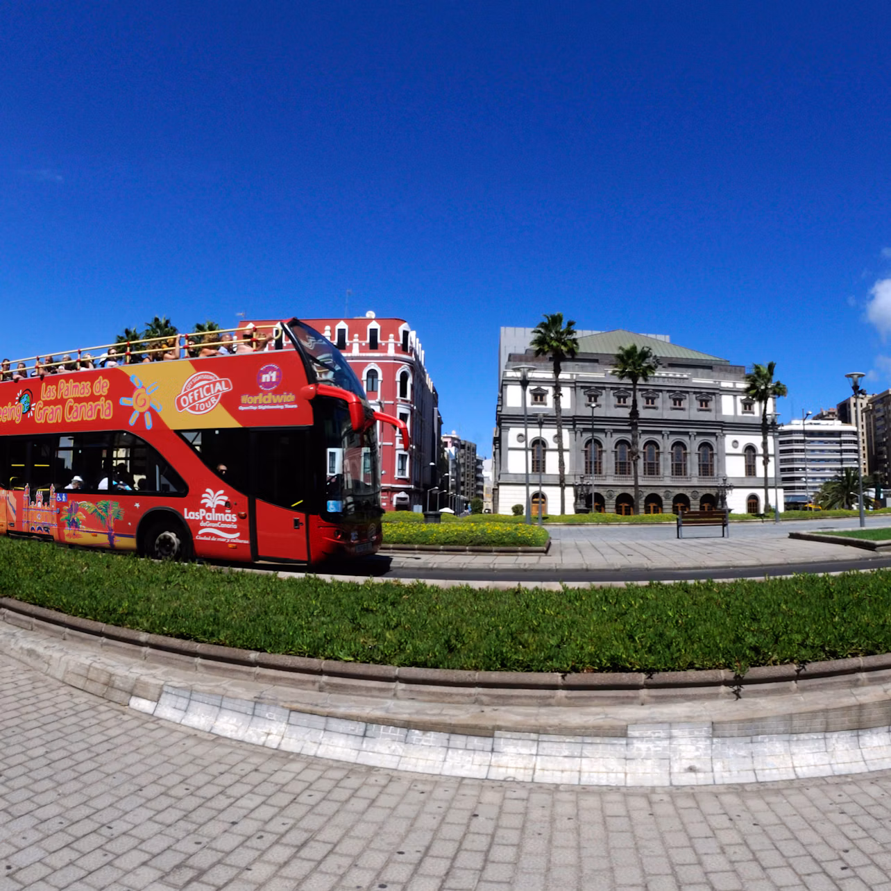 A red double-decker tour bus passes by a park with green grass, palm trees, and historical buildings under a bright blue sky.