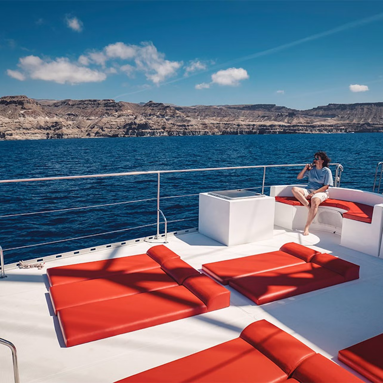 Person sitting on a boat deck with red cushions, overlooking calm blue water and rocky cliffs under a clear sky.