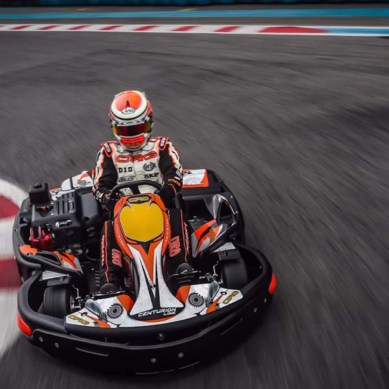 A person in racing gear and helmet drives a go-kart around a track corner. The go-kart has colorful decals and branding.