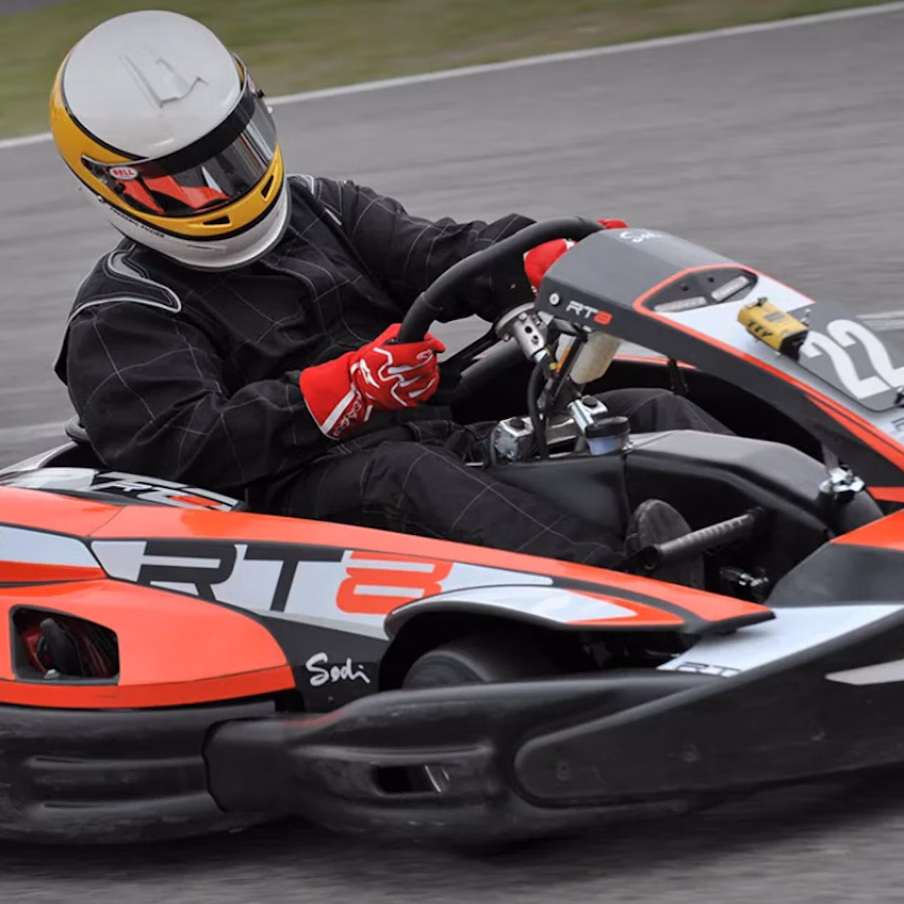 Person in a racing suit and helmet driving an orange and black go-kart on a track.