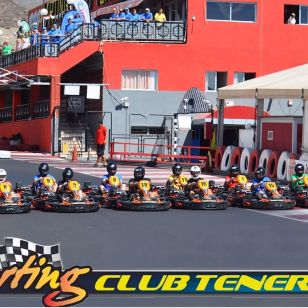 A group of go-karts lined up on a track in front of a red building with spectators above and a sign reading 'Club Tenerife.'