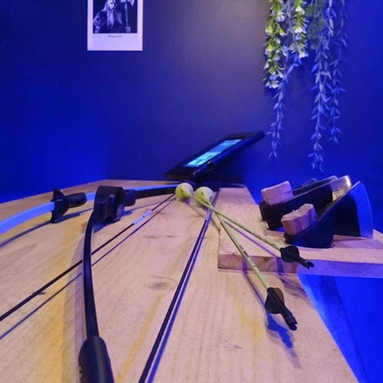 Close-up of a wooden table with two archery bows and a tablet. Wall in background has hanging plants and a framed photo.