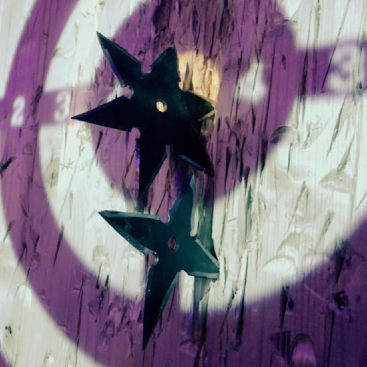 Two black throwing stars embedded in a wooden target board, illuminated by purple and white lights.