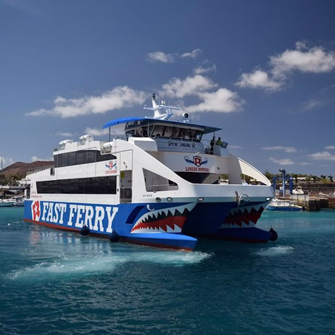 Large ferry boat labeled "FAST FERRY" on the side, sailing in clear blue water on a sunny day with a few clouds in the sky.