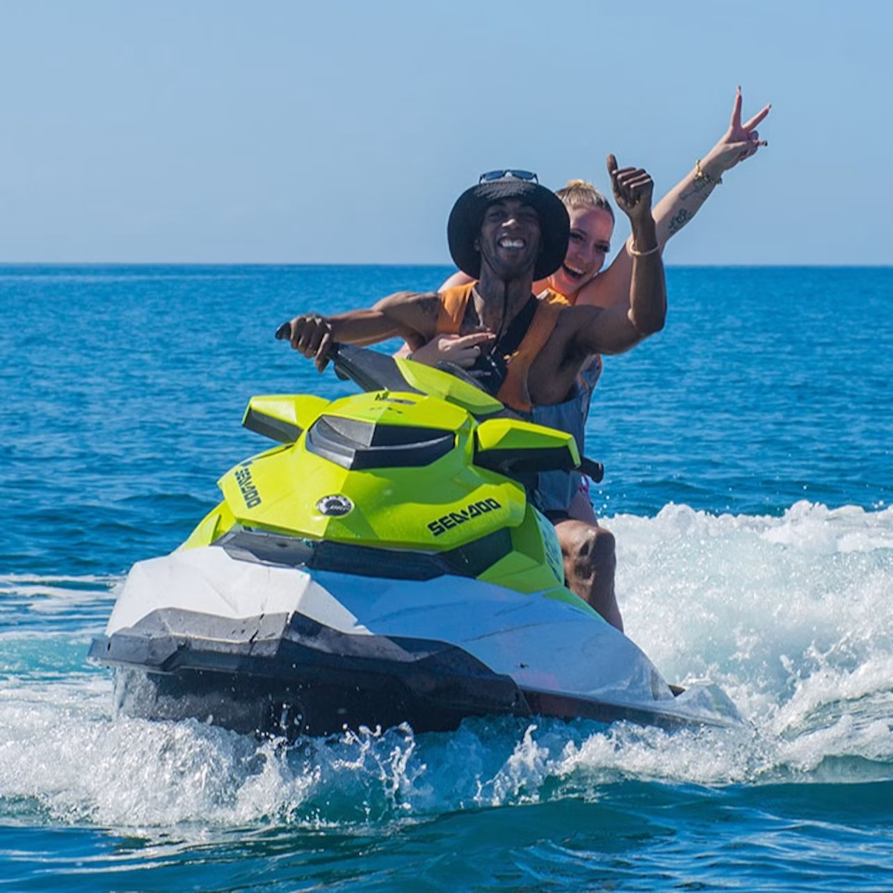 Two people on a yellow and white jet ski riding on blue water, with the passenger making a peace sign and the driver smiling.