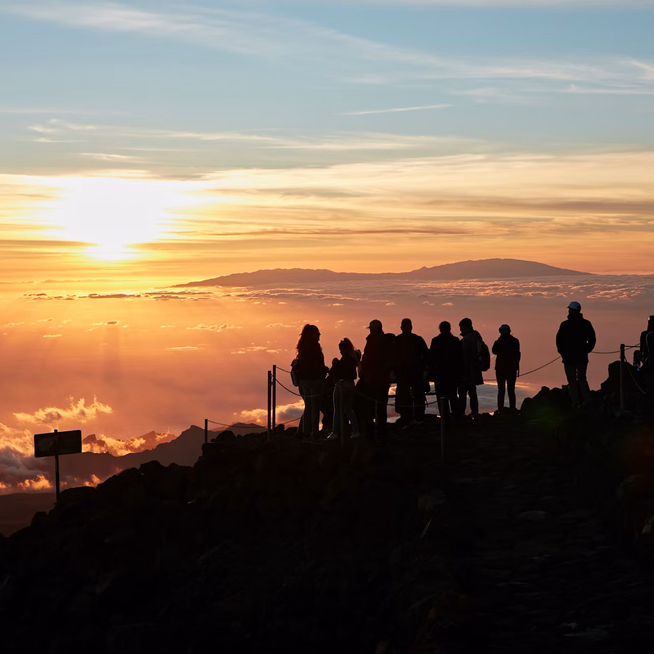 Silhouette of people standing on a rocky path at sunrise, overlooking a sea of clouds and distant mountains in the backdrop.