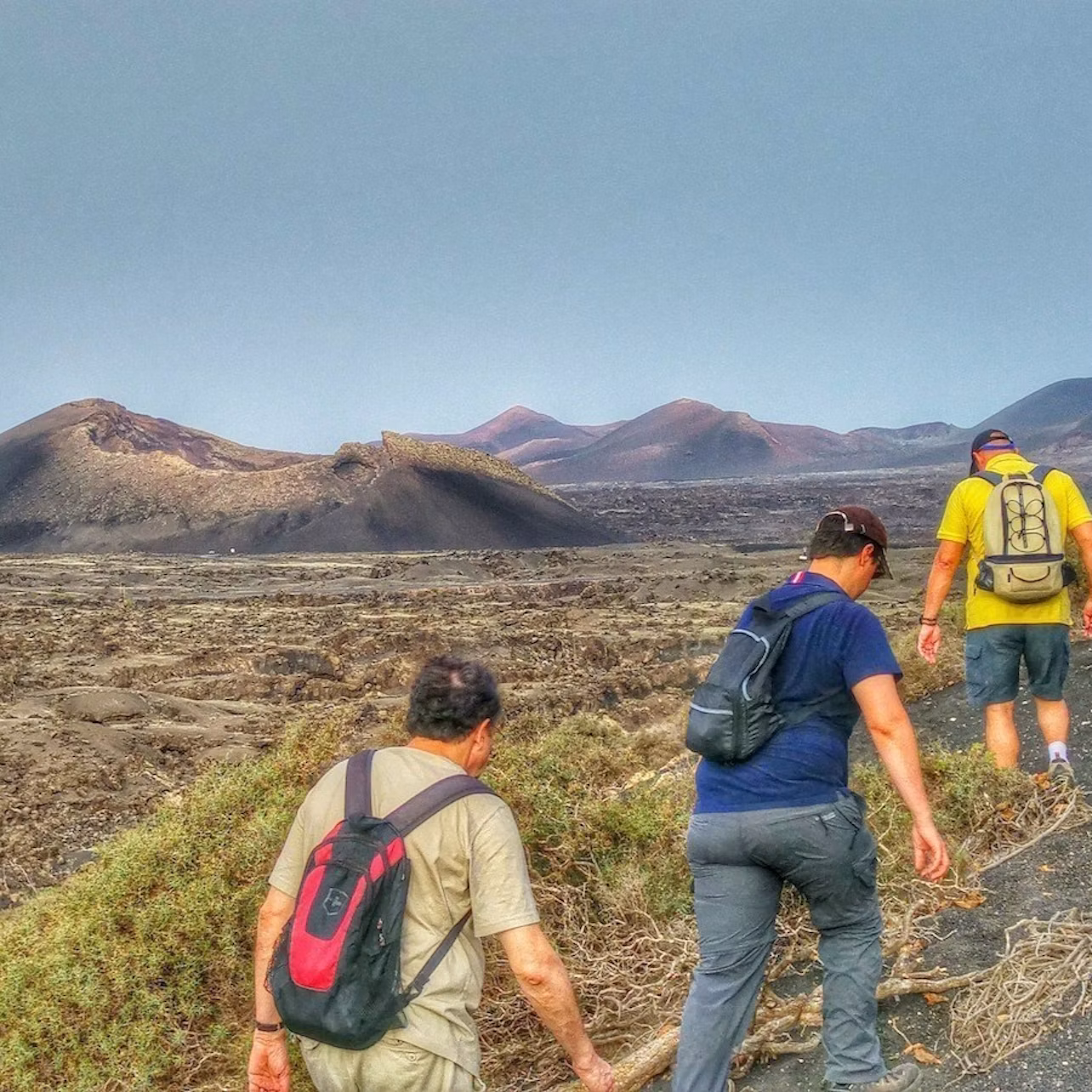 Three hikers with backpacks walking on a rocky terrain with volcanic hills and a clear sky in the background.