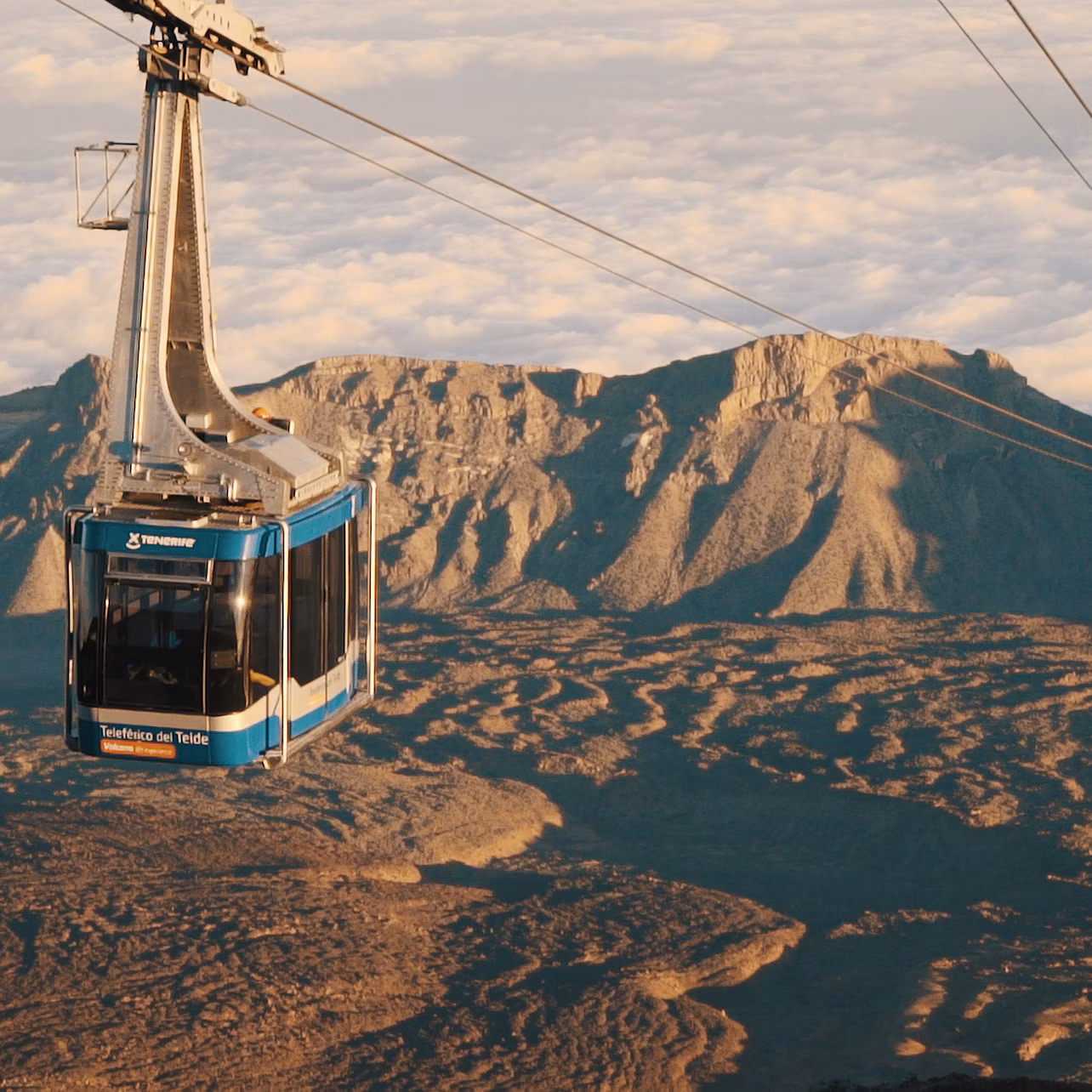 A blue cable car labeled "Teleférico del Teide" suspended over a rugged mountain landscape beneath a cloudy sky.