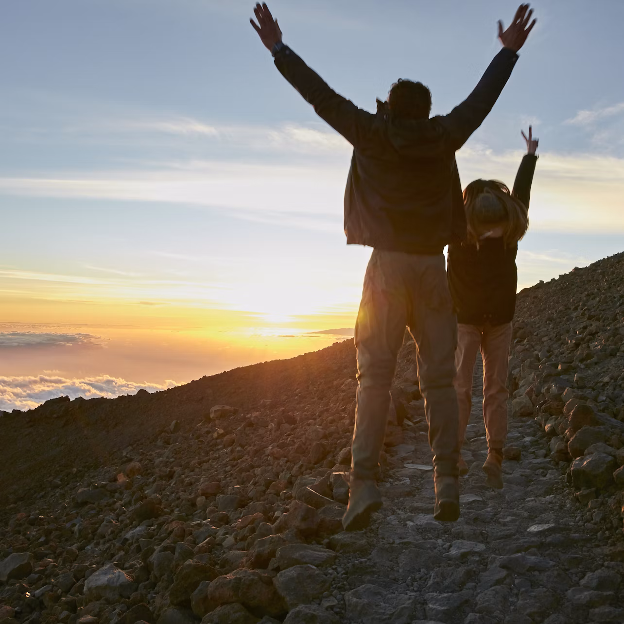 Two people with raised arms hike on a rocky path at sunset, with a scenic view of the sky and distant clouds.