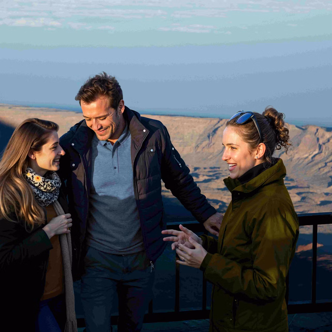 Three people in jackets talking and smiling near a railing overlooking a scenic, rocky landscape under a partly cloudy sky.