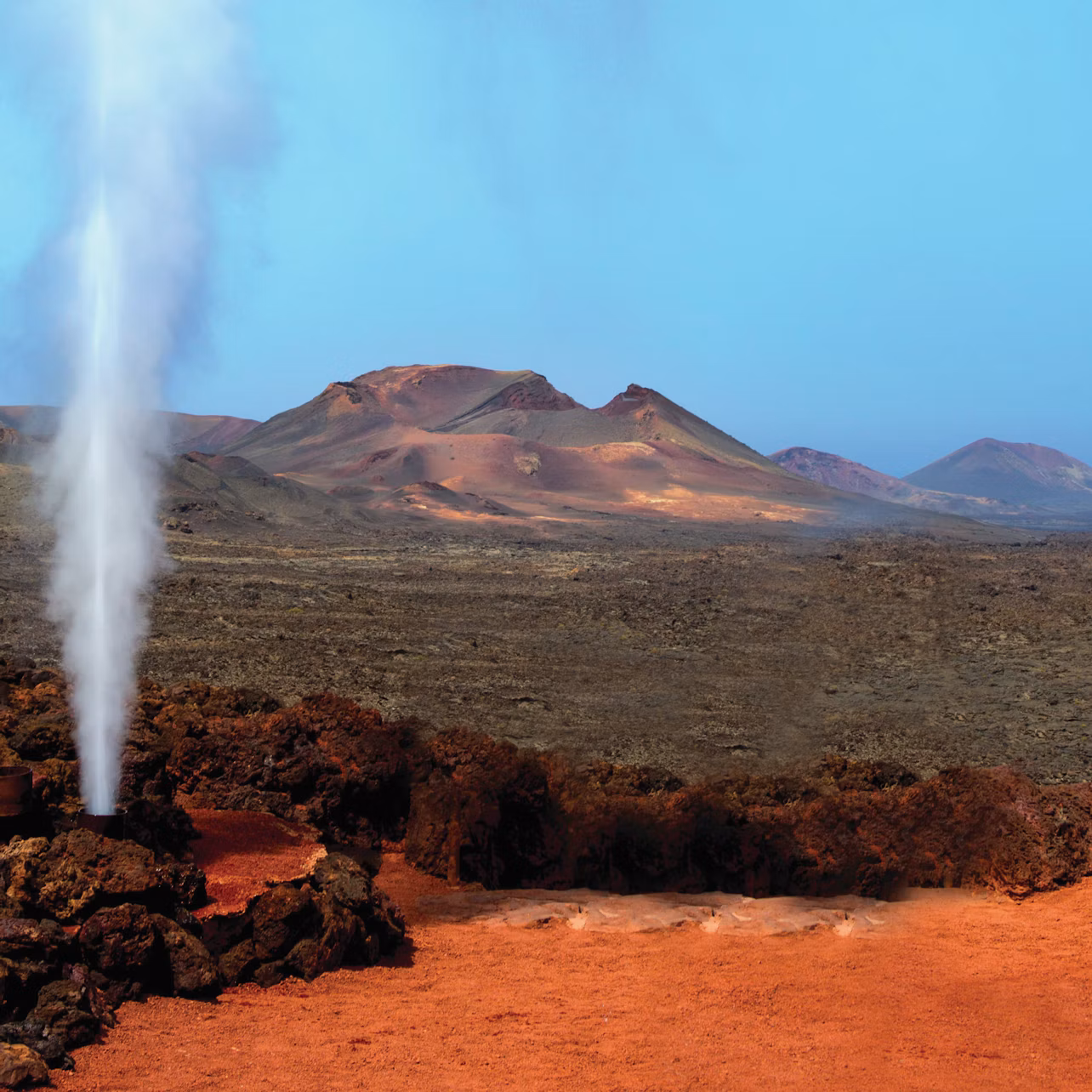 Steam geyser erupts from rocky terrain with red soil, set against a backdrop of barren, mountainous landscape under a clear blue sky.