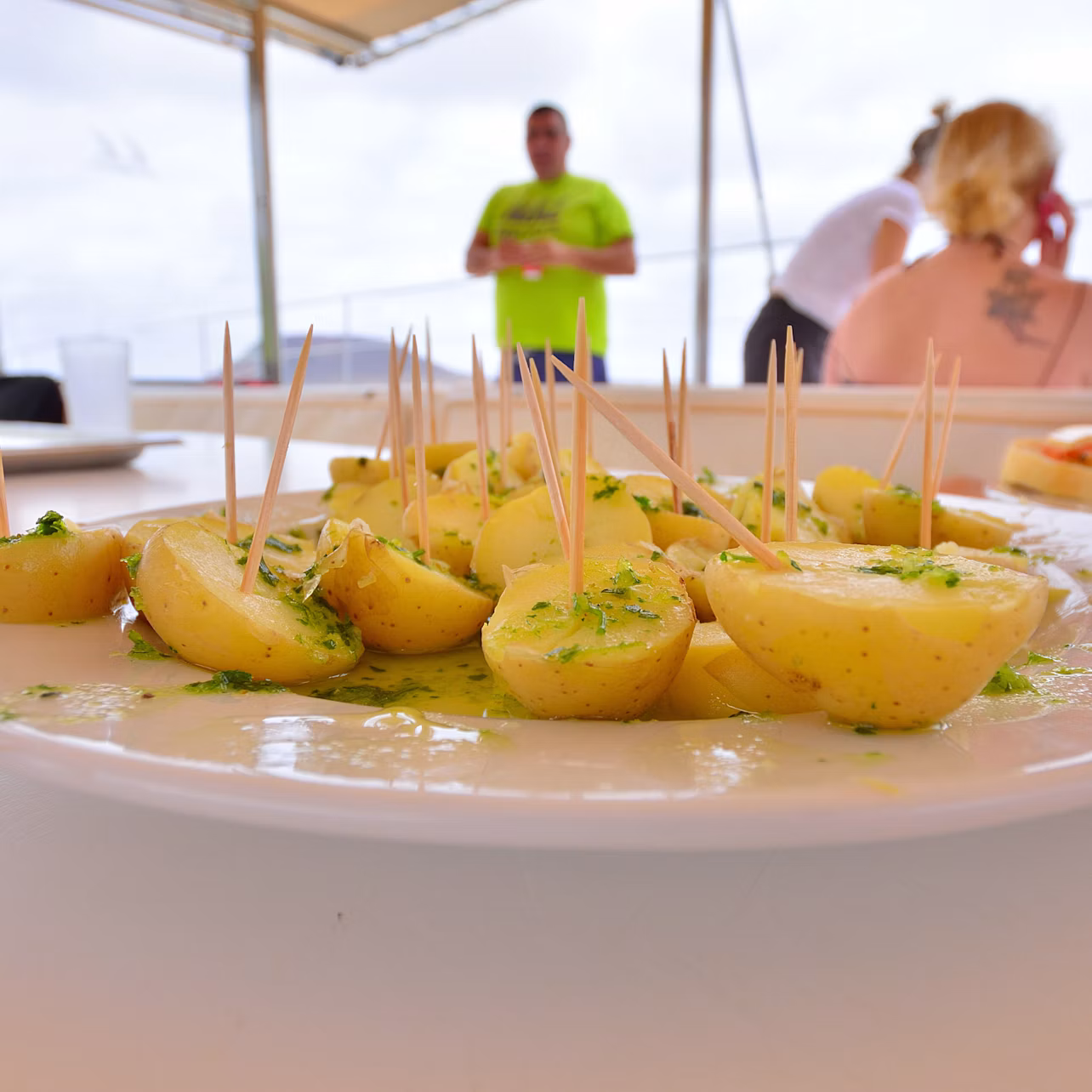 Plate of seasoned potato halves with toothpicks. Out-of-focus people and a bright outdoor setting in the background.