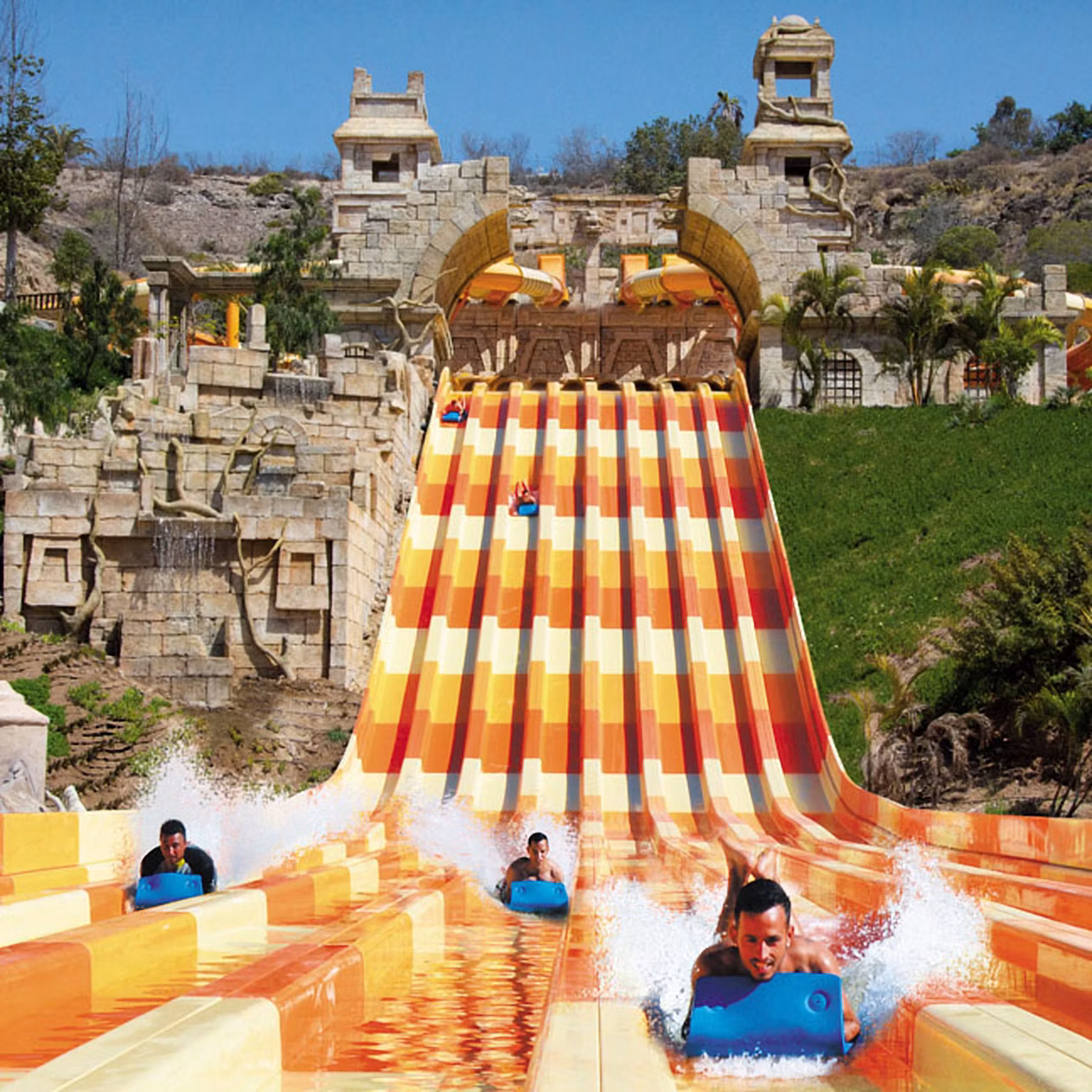 Four people slide down colorful water slides from a large, ornate structure resembling ancient ruins.