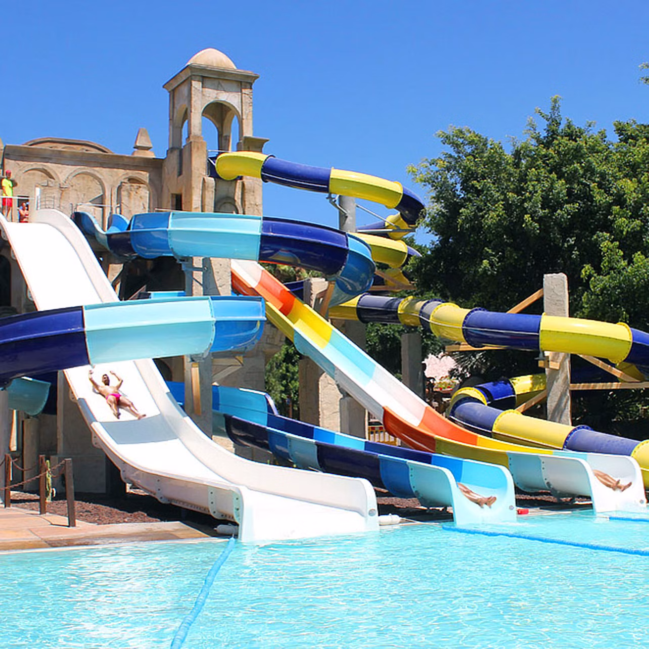 Multiple colorful water slides leading into a large pool with people sliding down, surrounded by trees and a building in the background.