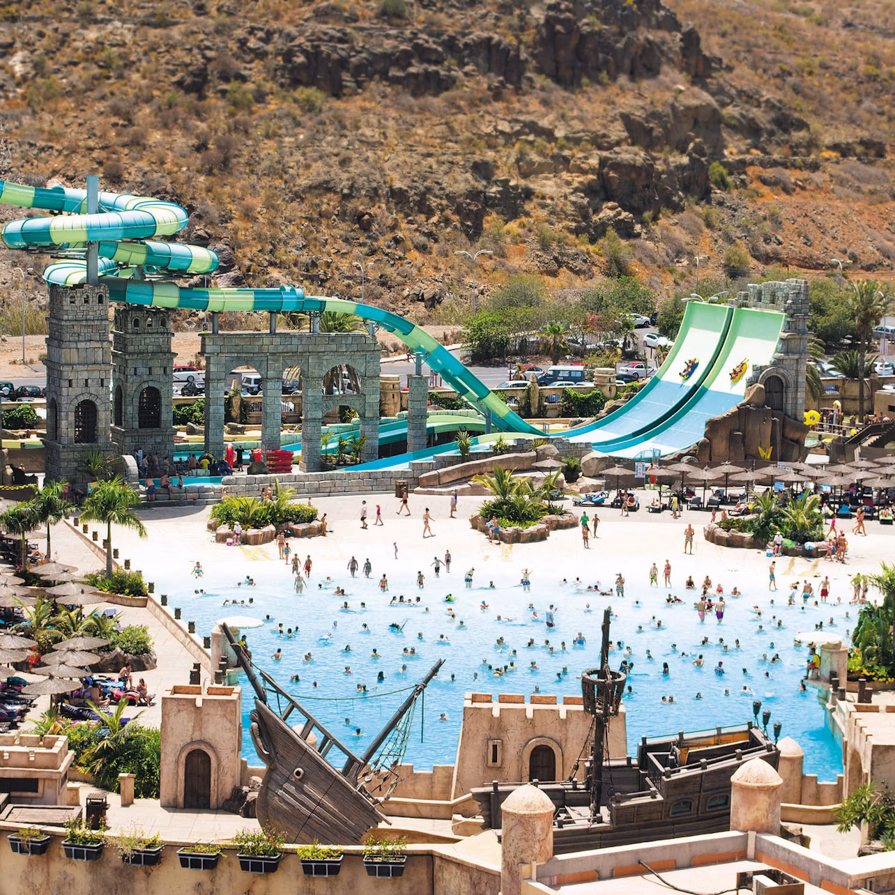 A water park with a wave pool, large slides, and a pirate ship structure surrounded by people enjoying the attractions.