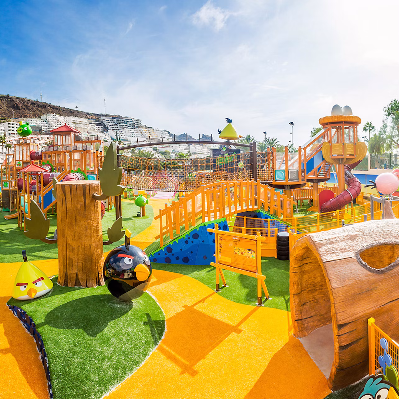Colourful playground with slides, bridges, climbing structures, and character-themed designs on a sunny day. Apartments visible in the background.