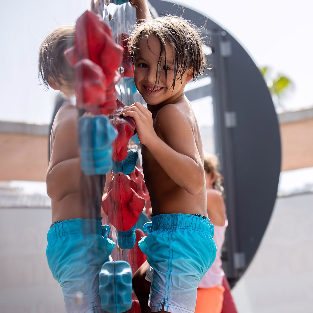 A child in blue shorts climbs on a playground structure, smiling towards the camera, with another child in the background.