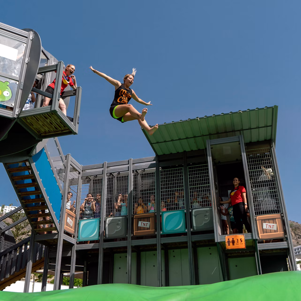 Person in mid-air jumping from a platform at an adventure park, with onlookers and a clear blue sky in the background.