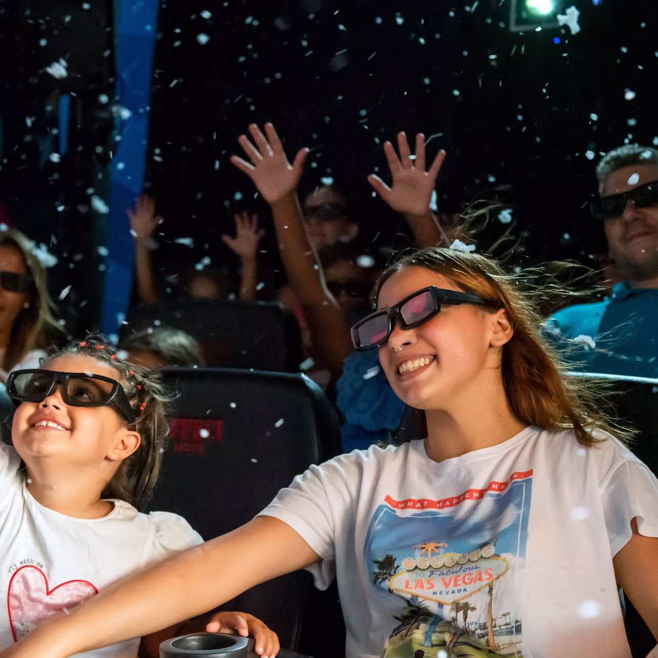 Children and adults wearing 3D glasses sit in a theatre, smiling and raising hands, with confetti or snow falling around them.