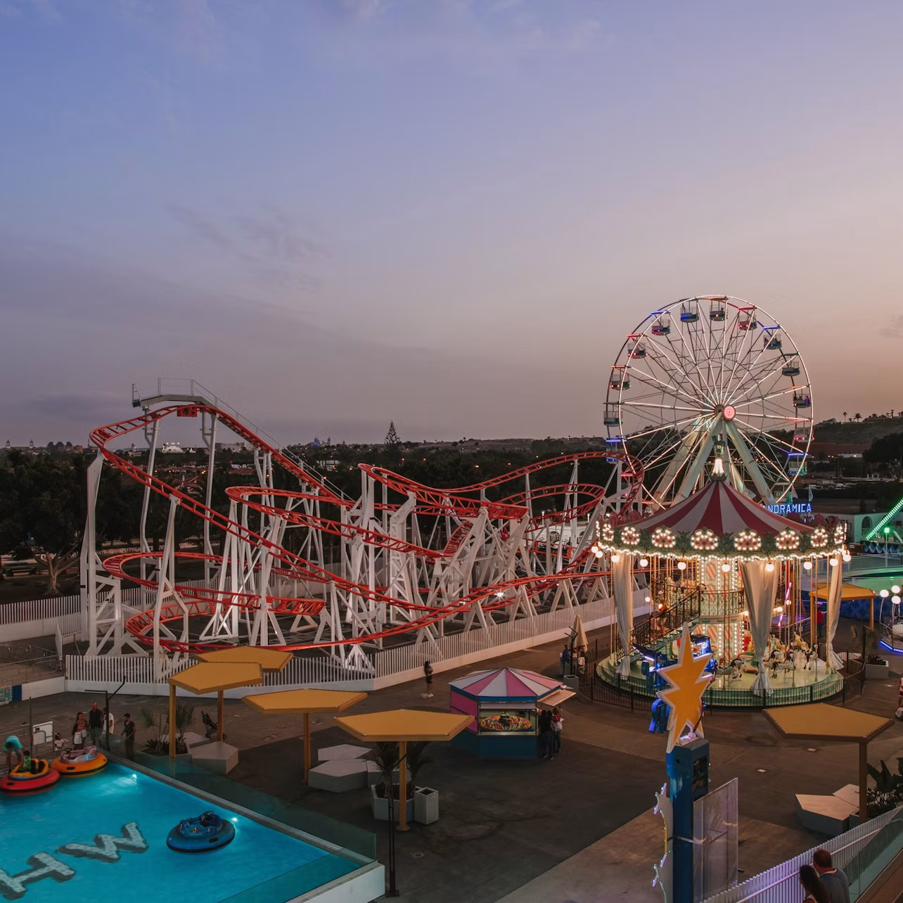 An amusement park at dusk with a rollercoaster, Ferris wheel, carousel, bumper boats, and colorful lights illuminating the area.