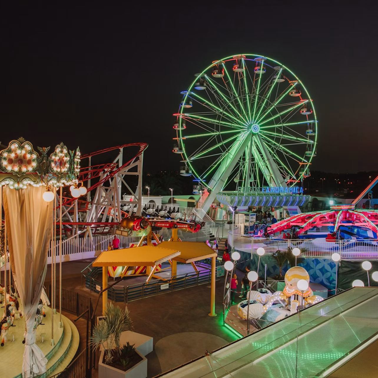 Amusement park at night with lit-up Ferris wheel, carousel, roller coasters, and colorful lights.