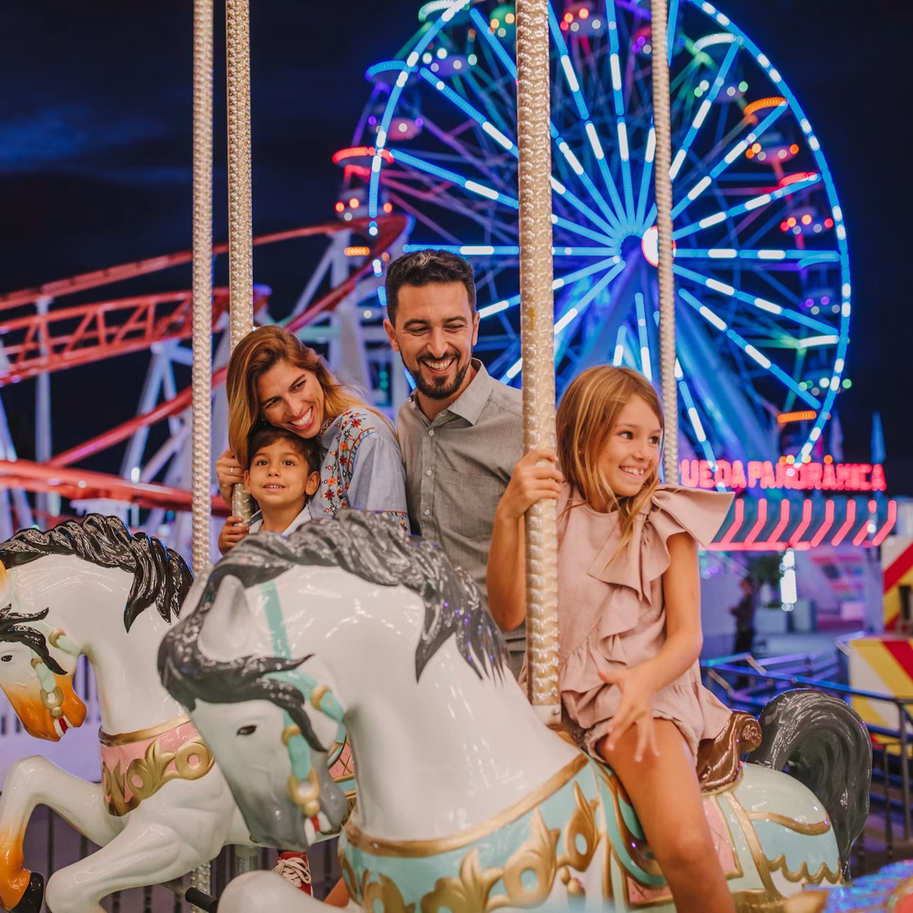 A family enjoying a carousel ride at night with a lit-up Ferris wheel and rollercoaster in the background.