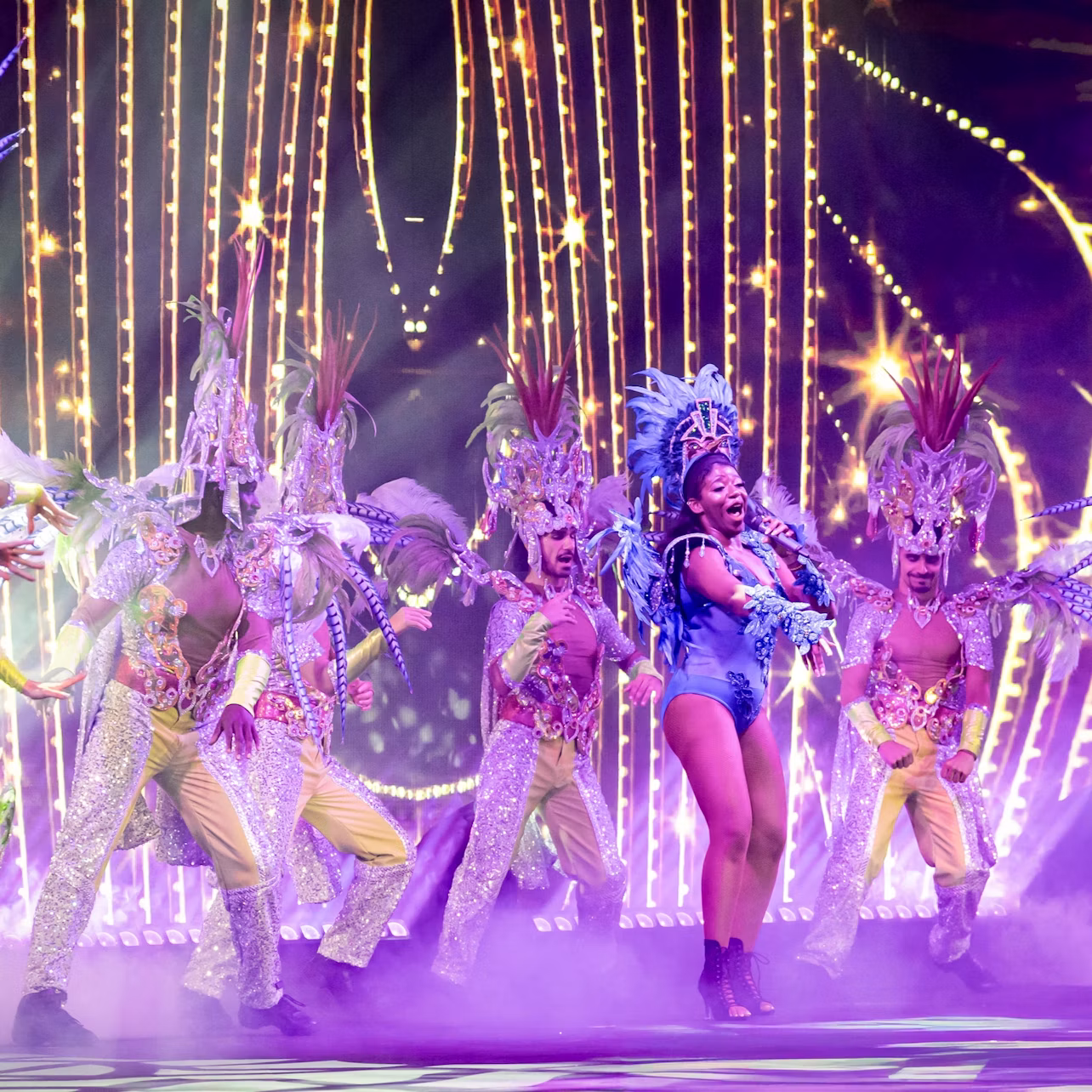A vibrant stage performance featuring dancers in elaborate costumes with bright colors and feathered headdresses.