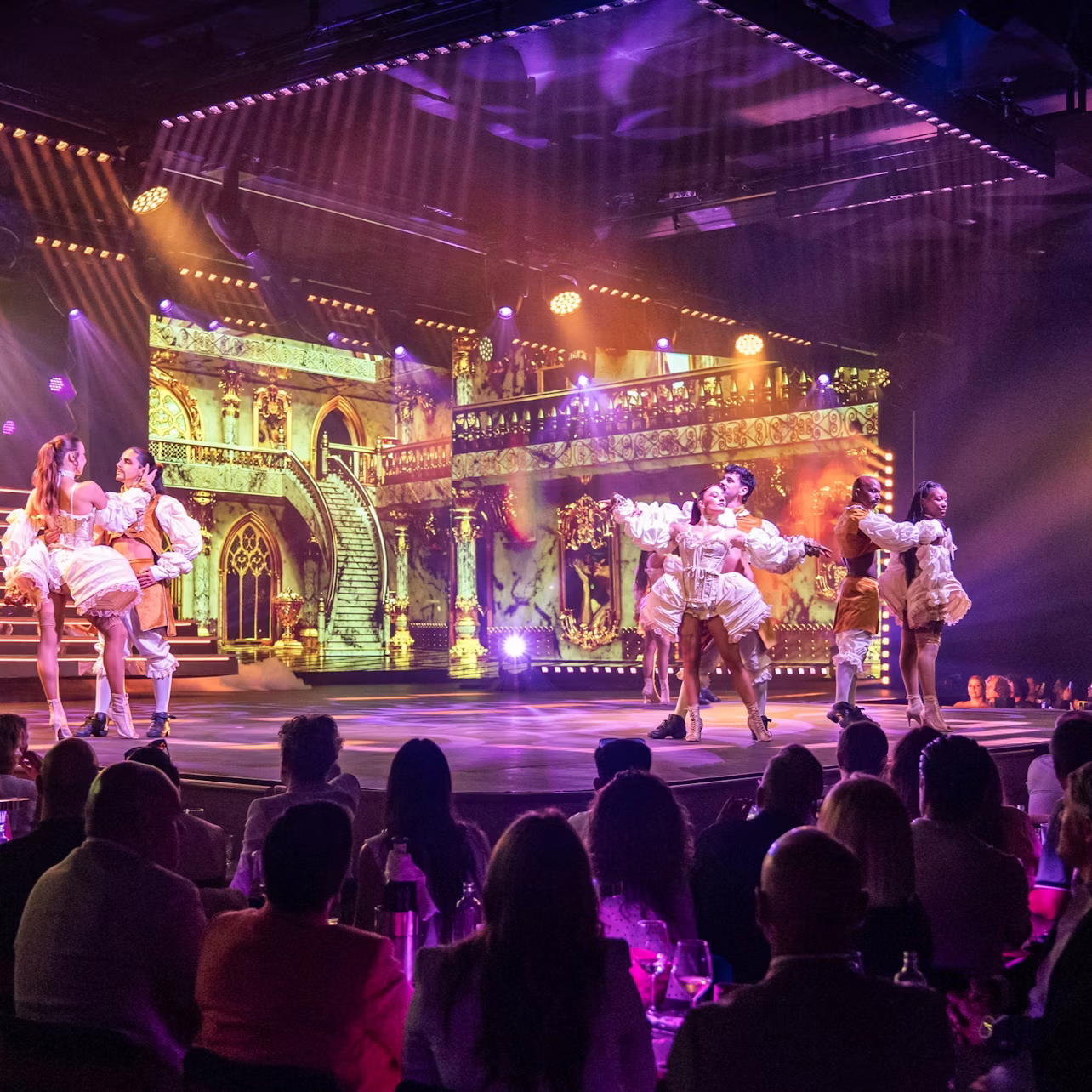 Performers in ornate costumes dance on a lit stage with a backdrop of a grand architectural scene, viewed by an audience.
