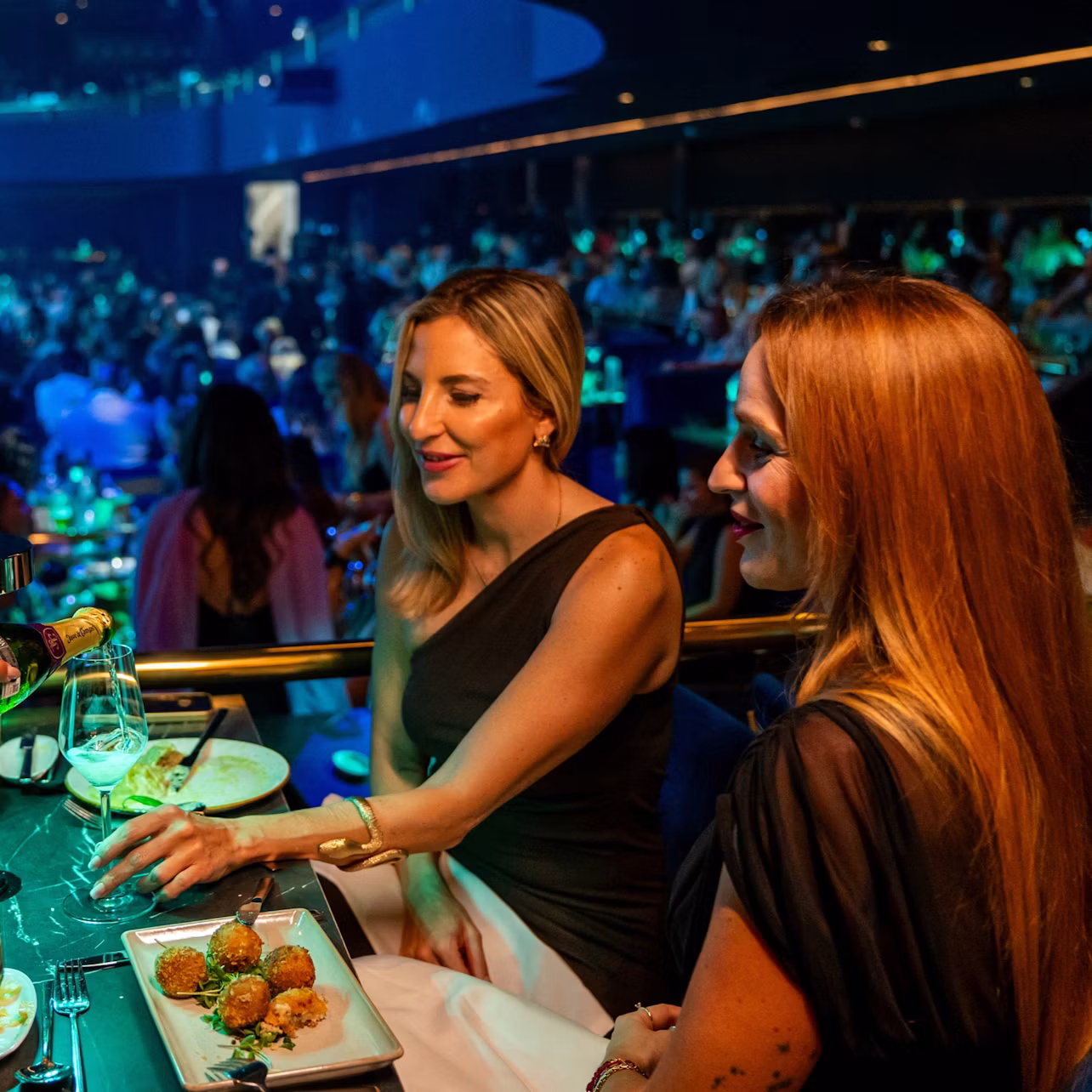 Two women sitting at a table with plates of food and drinks, in a lively, dimly lit venue filled with people.