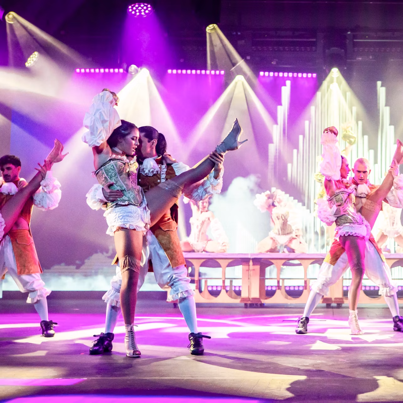 Performers in elaborate costumes dance on a brightly lit stage with colorful lights and a decorative backdrop.