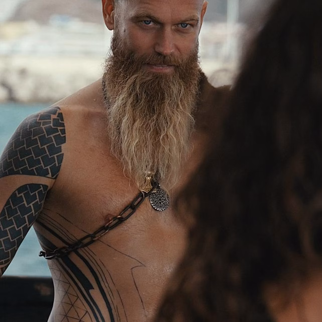 A man with a long beard, tattoos, and a chain necklace is facing a person with long curly hair, outdoors near water.