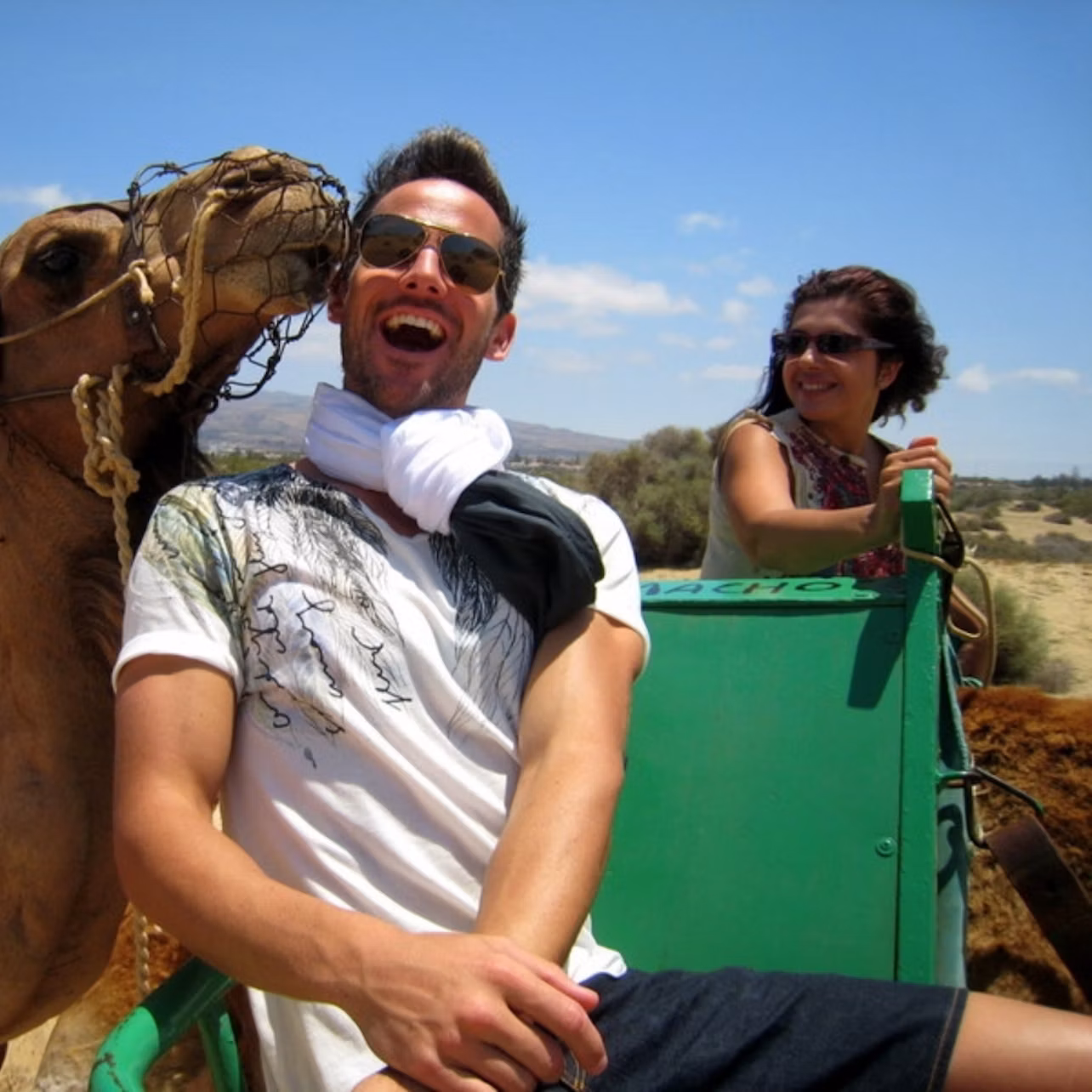 A man laughs while a nearby camel nuzzles his face. A woman smiles in the background, seated on a green cart.