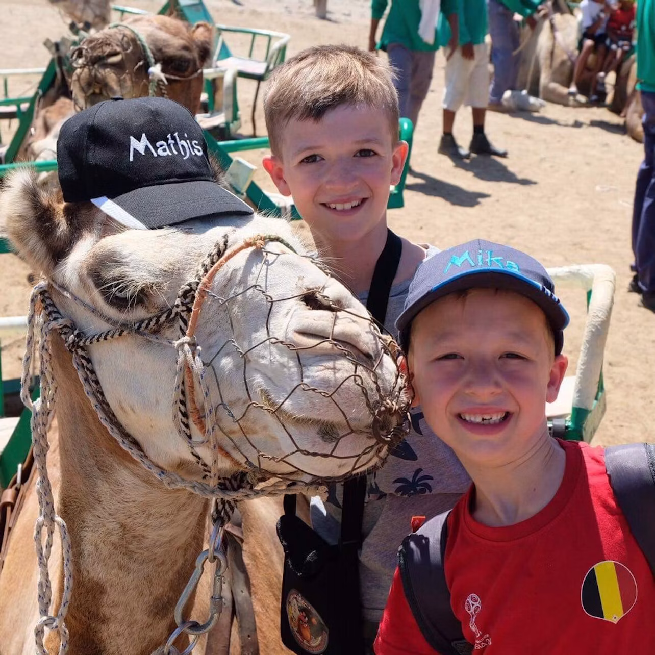Two boys pose next to a camel wearing a cap. The background shows other people and camels under a sunny sky.