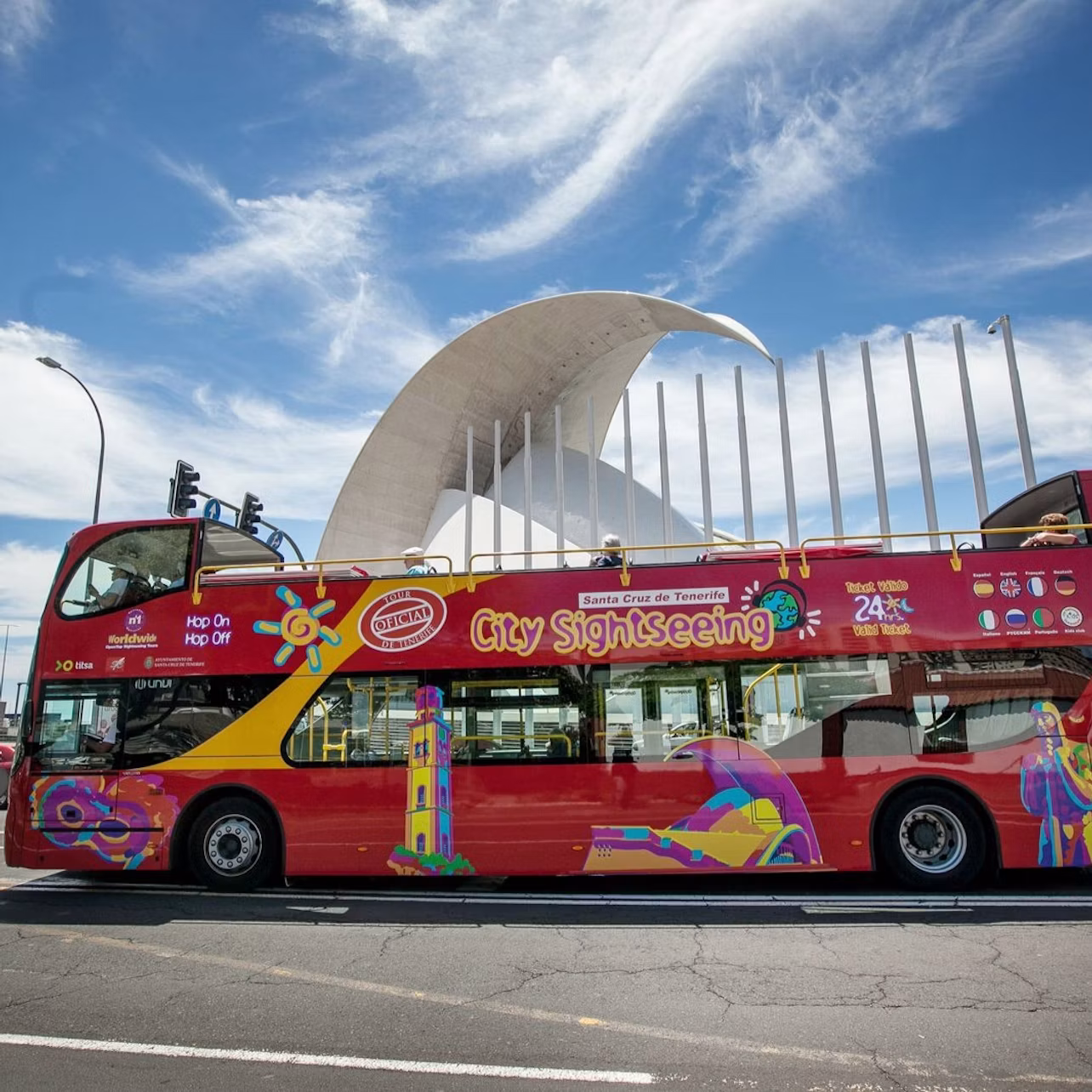 A red double-decker city sightseeing bus stands in front of a modern white architectural structure under a partly cloudy sky.
