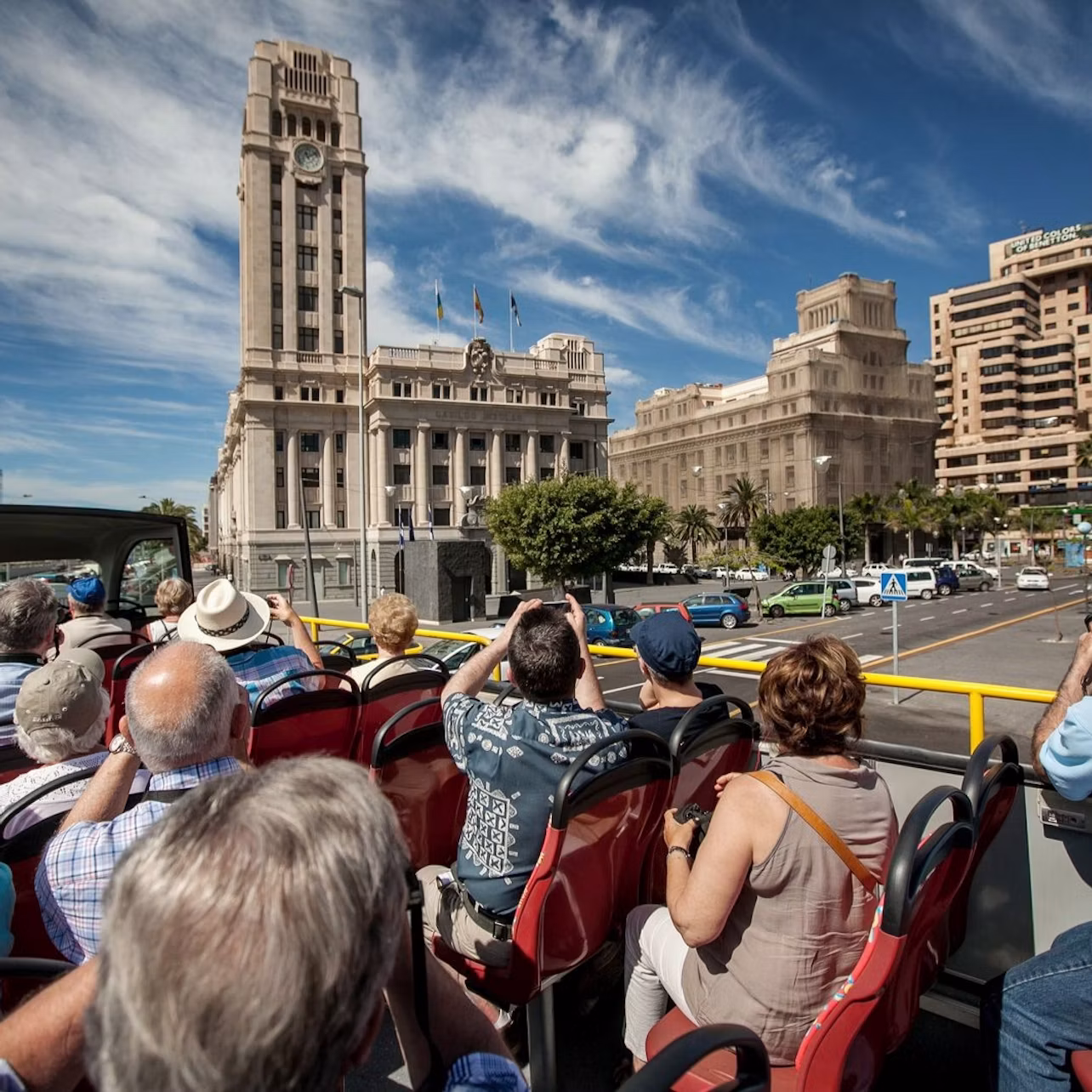 People seated on an open-top tour bus, taking photos of large historic buildings under a partly cloudy blue sky.