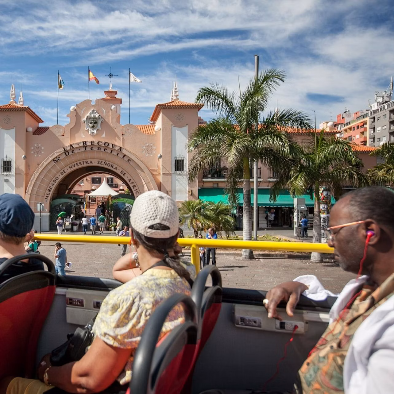 People on an open-top tour bus viewing a building entrance with flags, palm trees, and blue sky in the background.