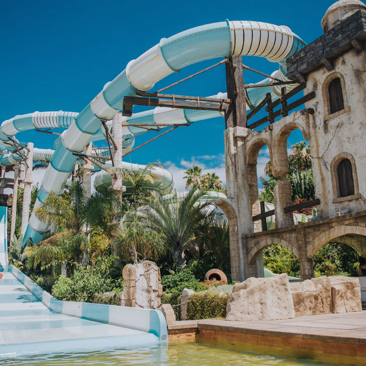 Outdoor water park with large blue and white tube slides, surrounded by palm trees and a stone structure, under a clear blue sky.