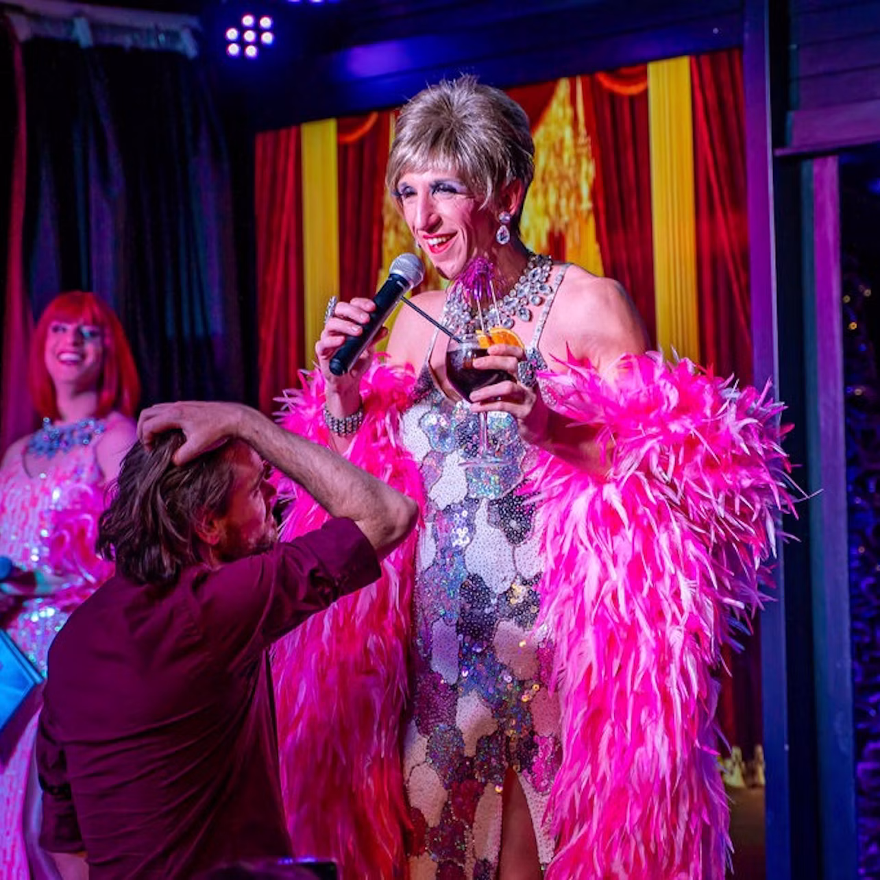 A performer in a sequined dress and pink feather boa holds a microphone and wine glass on stage, interacting with an audience member.