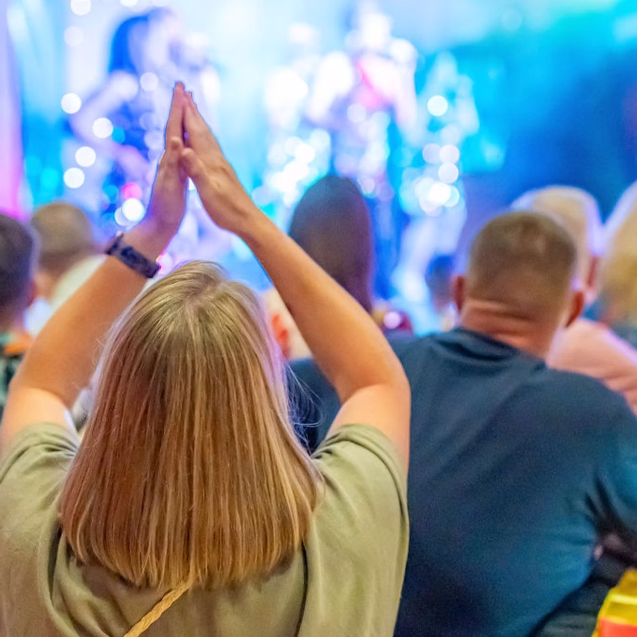A crowd watches a performance on stage, with one person raising their hands in the foreground.