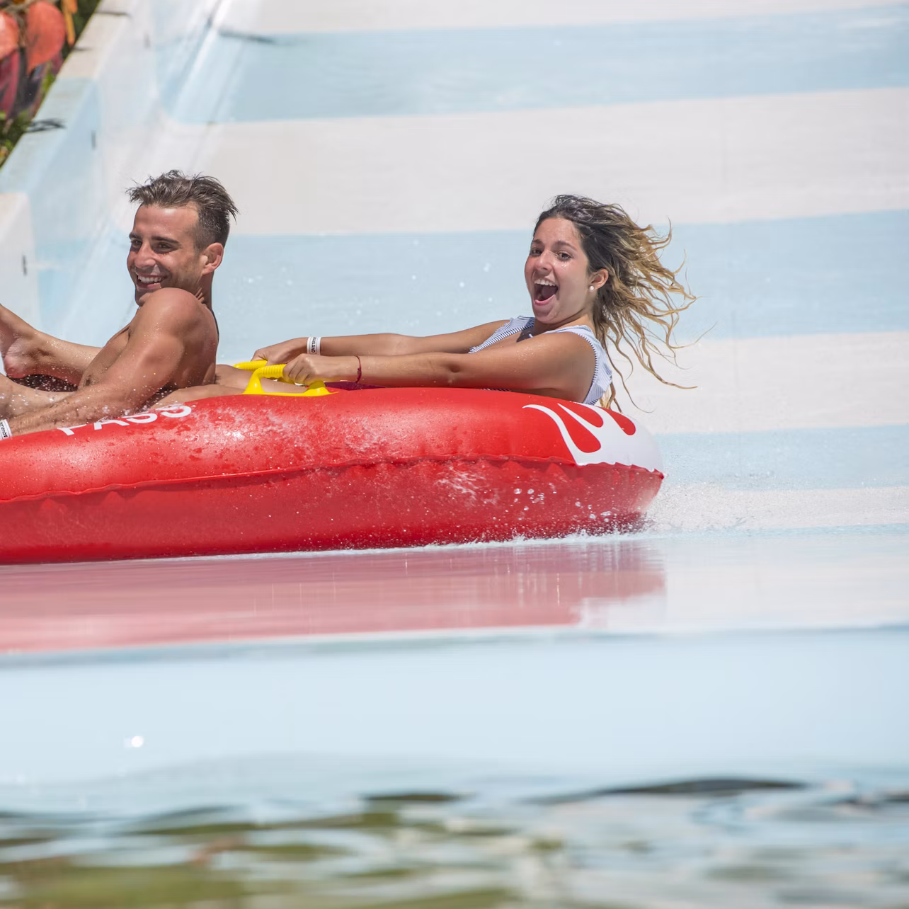 Two people excitedly riding a red inflatable raft down a water slide, splashing water around them.