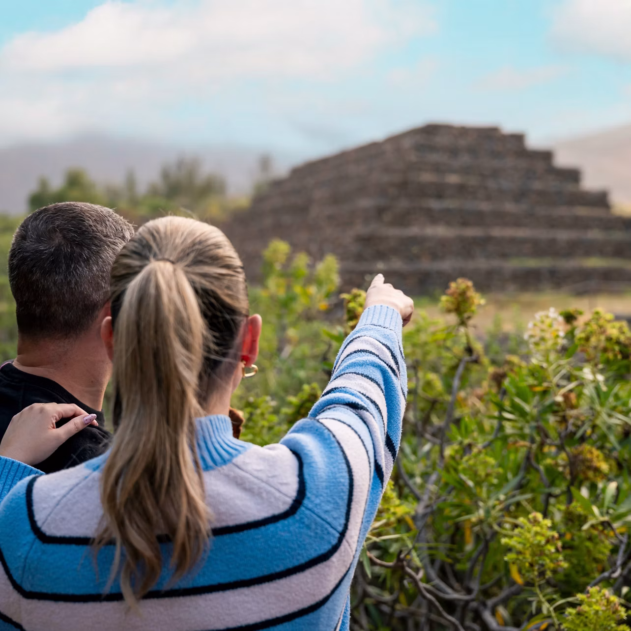A woman pointing at a stepped pyramid structure, standing next to a man, with vegetation in the foreground.