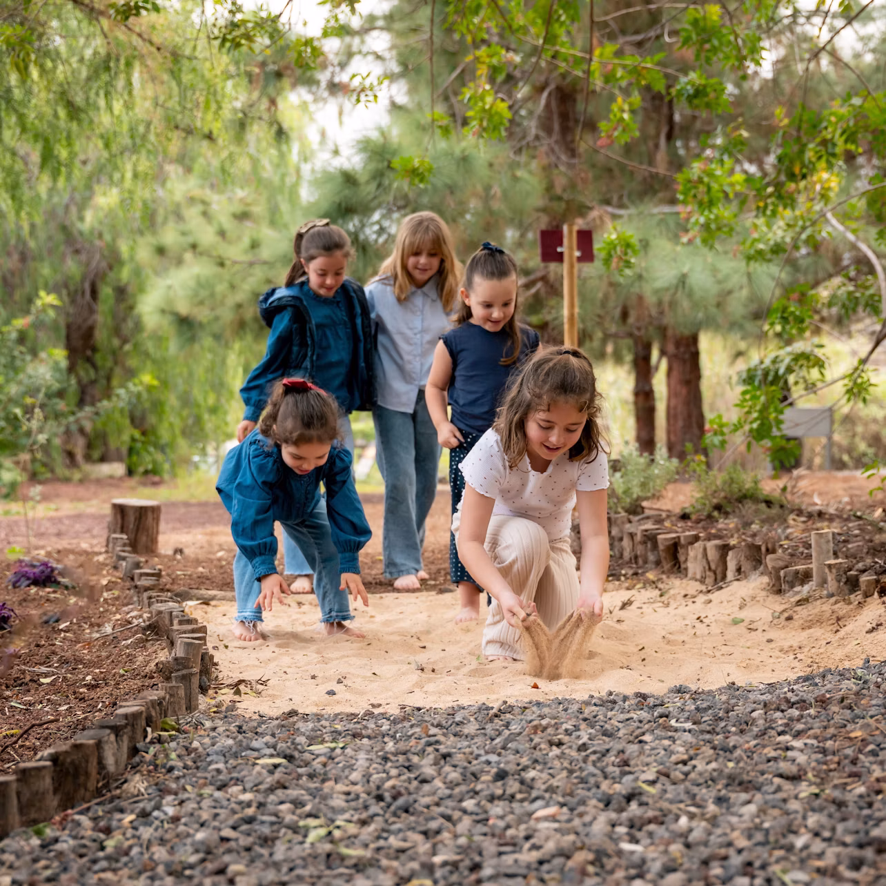 Five children playing on a sandy path in a wooded area, with trees and greenery surrounding them.