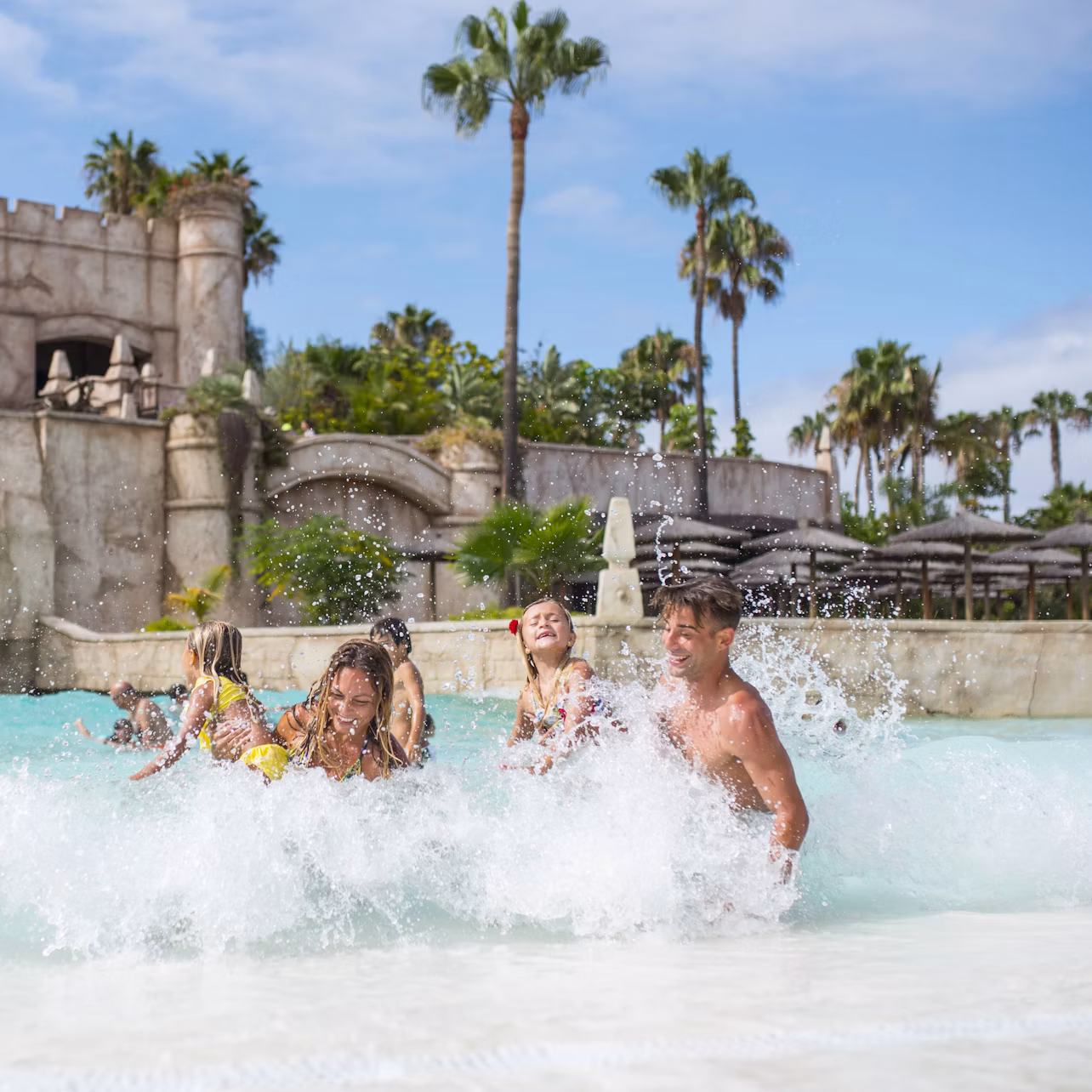 A family enjoying splashing in a shallow pool with a castle-like structure and palm trees in the background.