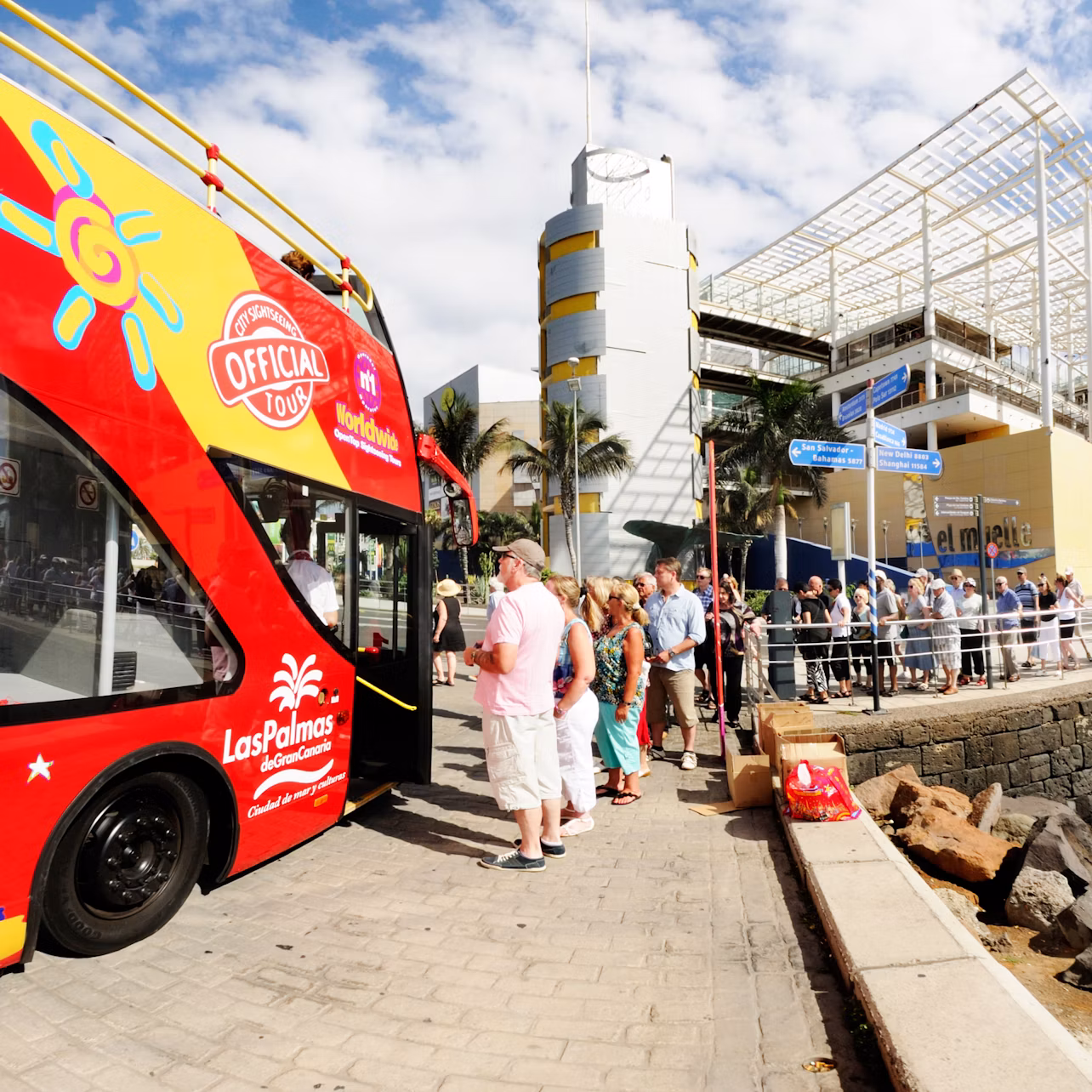 People stand in line to board a red double-decker bus labeled "Official Tour" near a modern building and informational signs.