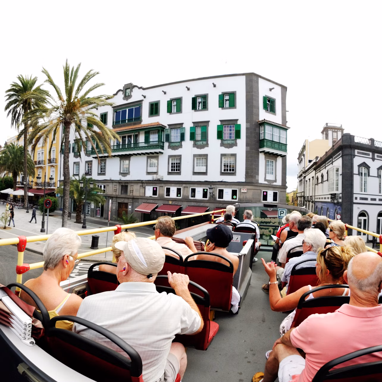 A group of tourists seated on the upper deck of an open-top bus, passing a building with green shutters and balconies in a sunny town.