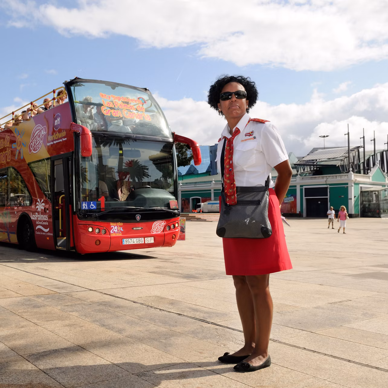 A tour guide stands in front of a red double-decker bus parked in an open area on a sunny day.