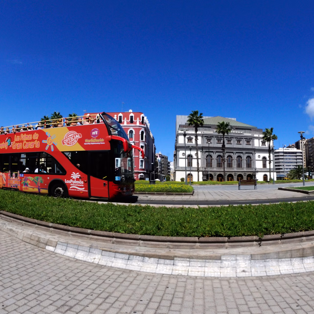 A red double-decker tour bus passes by a park with green grass, palm trees, and historical buildings under a bright blue sky.