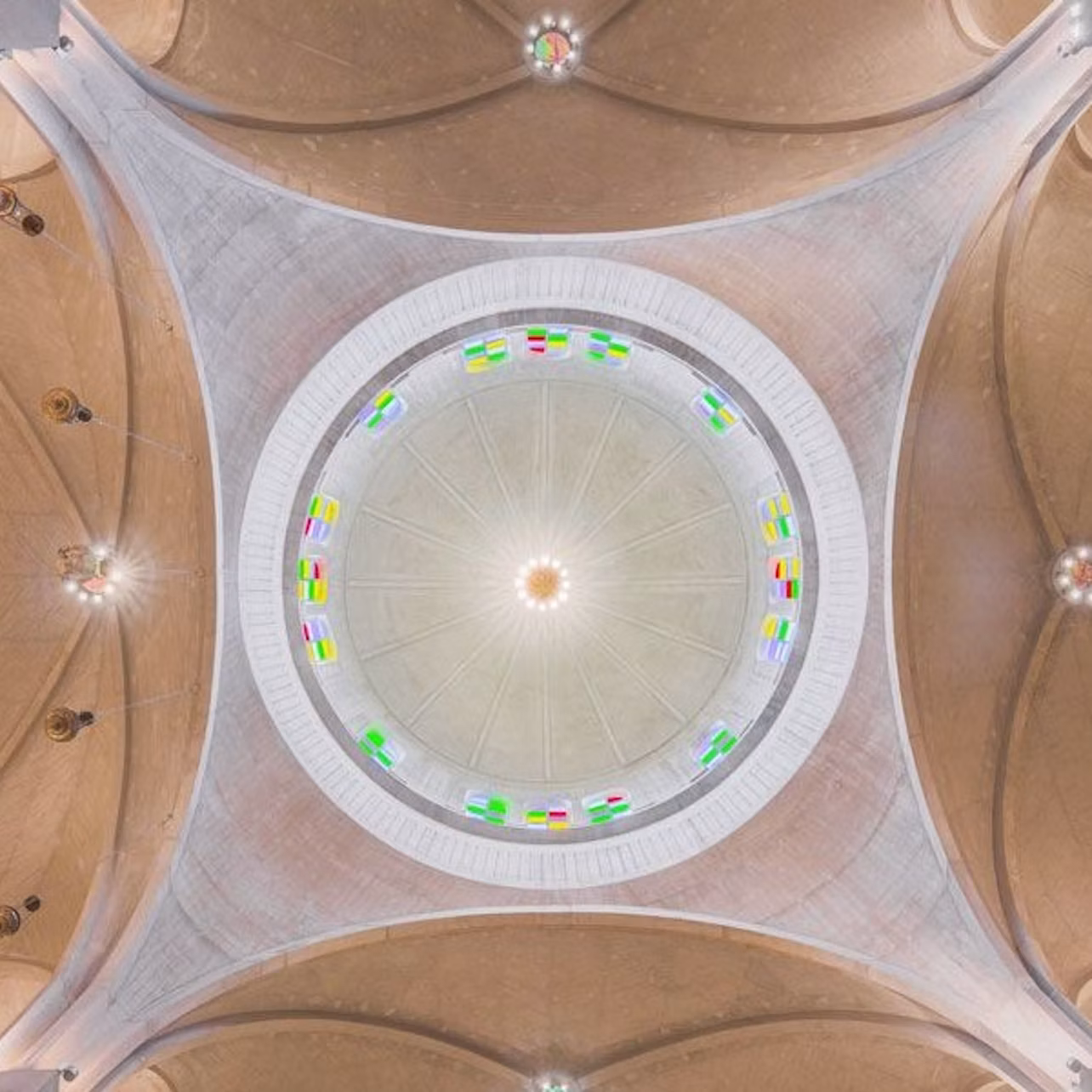 A symmetrical view of a dome ceiling with colorful stained glass windows encircling its base, set against a beige structure.